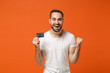 © ViDi Studio - Joyful happy young man in casual white t-shirt posing isolated on orange wall background, studio portrait. People lifestyle concept. Mock up copy space. Holding credit bank card, doing winner gesture.