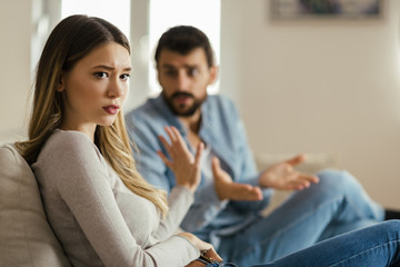 Wall Mural - Worried young woman sitting on sofa at home and ignoring her boyfriend who is sitting next to her