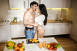 Young sexy couple have intimacy in kitchen in night. Beautiful hot woman embrace and kiss shirtless guy. Model wear white shirt and lingerie. Fruit and vegetables on table.
