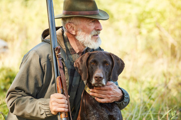 Caucasian mature man with gun and dog sit searching prey. Bearded man in hunting clothes. Autumn