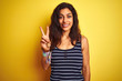 Young beautiful woman wearing striped t-shirt standing over isolated yellow background showing and pointing up with fingers number two while smiling confident and happy.