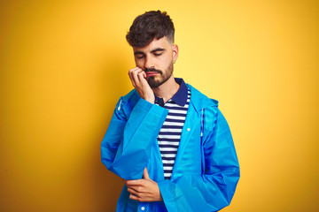 Wall Mural - Young handsome man wearing rain coat standing over isolated yellow background thinking looking tired and bored with depression problems with crossed arms.