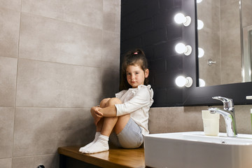 brunette girl, in white pullover, shorts and socks, sitting near clean sink with serious face, looks streight expressing confidence, spreds hands round bend legs, portrait, shot from below