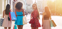 Just Turn Around. A Multicultural Group Of Four Girls Walking Along The City Streets With Shopping Bags, Turned Their Backs To The Camera, While One Brunette In Red Dress Turns Around And Smiles.