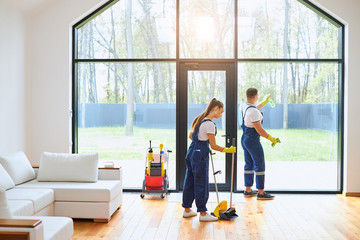 Young cleaners in blue uniform mopping wooden floor, cleaning big panoramic window, preparing country house for selling. Cleaning service concept