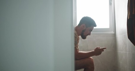 in the bathroom young man on toilet relaxing playing mobile game on smartphone winning cheering up. 
