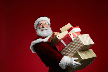 Waist up portrait of smiling Santa Claus holding stack of Christmas presents over red background, copy space