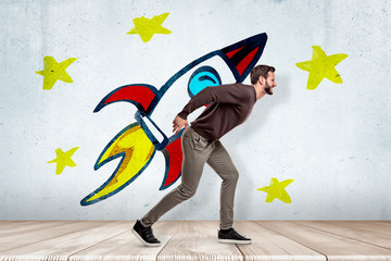 Young man in casual clothes carrying heavy colorful cartoon space rocket on his back on white wall with yellow stars background