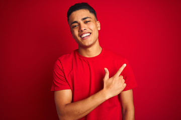 Wall Mural - Young brazilian man wearing t-shirt standing over isolated red background cheerful with a smile on face pointing with hand and finger up to the side with happy and natural expression