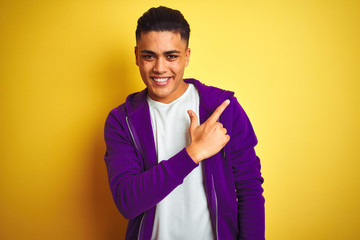 Young brazilian man wearing purple sweatshirt standing over isolated yellow background cheerful with a smile on face pointing with hand and finger up to the side with happy and natural expression