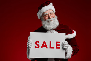 Wall Mural - Waist up portrait of classic Santa Claus holding SALE sign and smiling while standing against red background, copy space