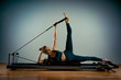 © Georgii - Young girl doing pilates exercises with a reformer bed. Beautiful slim fitness trainer on reformer gray background, low key, art light. Fitness concept