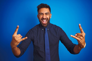 Wall Mural - Young indian businessman wearing elegant shirt and tie standing over isolated blue background shouting with crazy expression doing rock symbol with hands up. Music star. Heavy concept.