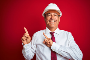 Wall Mural - Handsome middle age engineer man wearing security helmet over isolated red background smiling and looking at the camera pointing with two hands and fingers to the side.