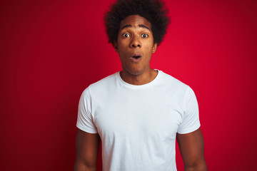 Poster - Young american man with afro hair wearing white t-shirt standing over isolated red background afraid and shocked with surprise expression, fear and excited face.