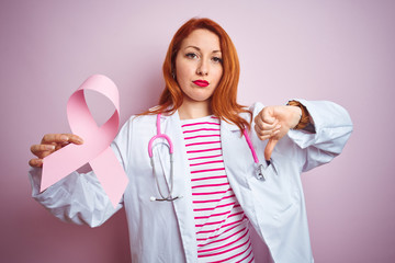 Sticker - Young redhead doctor woman holding cancer ribbon over pink isolated background with angry face, negative sign showing dislike with thumbs down, rejection concept