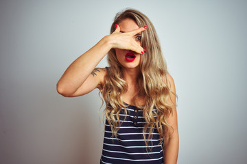 Sticker - Young beautiful woman wearing stripes t-shirt standing over white isolated background peeking in shock covering face and eyes with hand, looking through fingers with embarrassed expression.