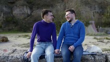 Two Gay Guys From The LGBT Community, Sitting On A Stone Pier In A Sweater And Sweatshirt And Scrubbing While Holding Each Other's Hands