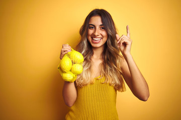 Sticker - Young beautiful woman holding net of lemons over yellow isolated background very happy pointing with hand and finger to the side