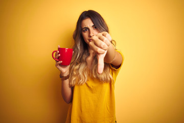 Sticker - Young beautiful woman holding  red cup of coffee over yellow isolated background with angry face, negative sign showing dislike with thumbs down, rejection concept