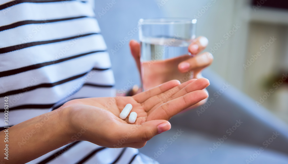 Woman holding white pill on hand and drinking water in glass on sofa in house, feels like sick. Health care concept. - obrazy, fototapety, plakaty 