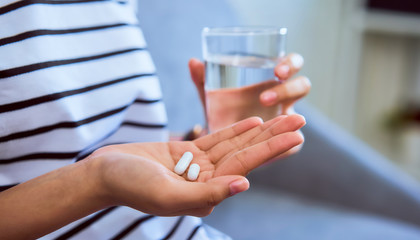 woman holding white pill on hand and drinking water in glass on sofa in house, feels like sick. heal