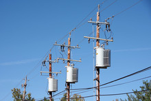 Low Angle Upward View Of A Row Of Tall Electricity Distribution Poles With Transformers And Power Lines Seen Against A Clear Blue Sky