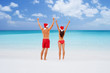 Christmas december travel fun couple on beach holiday wearing santa hats with arms raised up in happiness wearing swimwear walking relaxing on Caribbean tropical vacation destination for Xmas.