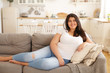 Portrait of positive young overweight brunette female sitting barefooted on gray sofa with legs outstretched wearing ripped jeans and white t-shirt looking at camera with happy cheerful smile