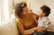 © shurkin_son - Indoor shot of happy young Hispanic woman with brown wavy hair relaxing at home embracing her adorable toddler son. Cheerful mother bonding with infant son, sitting on sofa in living room, laughing