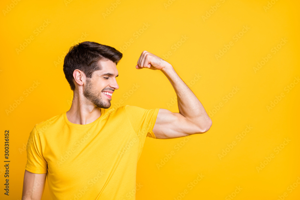 Photo of young handsome guy holding fist raised demonstrating perfect big biceps result after new sport gym wear casual t-shirt isolated yellow color background - obrazy, fototapety, plakaty 