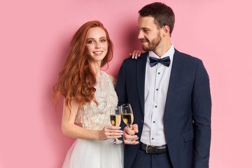 Wall Mural - Caucasian nice-looking lovely cheerful couple celebrating their marriage drinking champagne isolated over pink background. Love, celebration concept. Man in tuxedo look with love at woman in dress