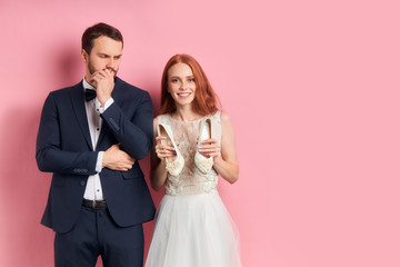 Wall Mural - Portrait of positive redhaired woman in suit seriously looking at smiling female in white wedding dress. isolated over pink background