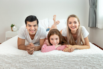 Happy family, including father mother and daughter, are lying down on bed with smile, looking at camera