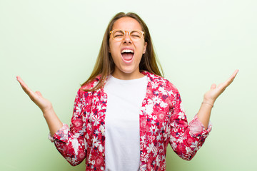 Wall Mural - young woman furiously screaming, feeling stressed and annoyed with hands up in the air saying why me against green background