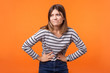 © khosrork - Portrait of unhappy ill woman with brown hair in long sleeve striped shirt standing, holding her belly with hands, stomach cramps or period pain. indoor studio shot isolated on orange background