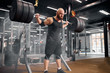 Brutal rude man powerlifter standing in the middle of big brightly lighted gym room, holding barbell, trying to raise above head, expressing power and strength, indoor shot