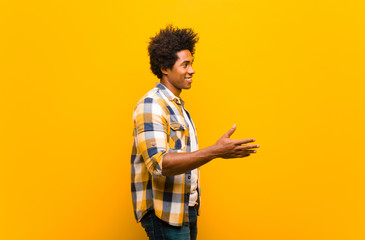 Wall Mural - young black man smiling, greeting you and offering a hand shake to close a successful deal, cooperation concept against orange wall