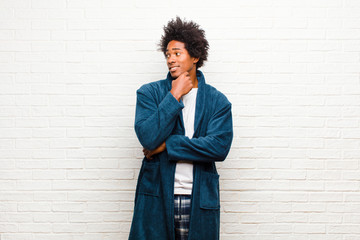 Wall Mural - young black man wearing pajamas with gown smiling with a happy, confident expression with hand on chin, wondering and looking to the side against brick wall