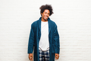 Wall Mural - young black man wearing pajamas with gown looking happy and friendly, smiling and winking an eye at you with a positive attitude against brick wall