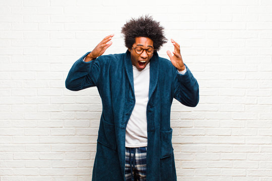 young black man wearing pajamas with gown screaming in panic or anger, shocked, terrified or furious, with hands next to head against brick wall