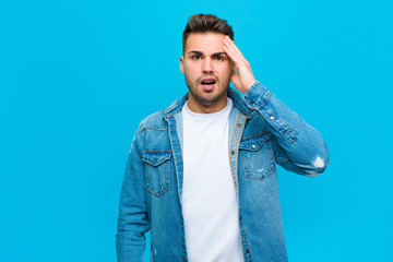 Wall Mural - young hispanic man looking happy, astonished and surprised, smiling and realizing amazing and incredible good news against blue background