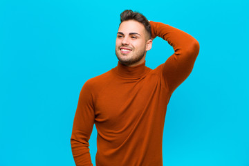 Wall Mural - young hispanic man smiling cheerfully and casually, taking hand to head with a positive, happy and confident look against blue background