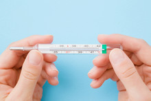 Young Woman Hands Holding Mercury Thermometer On Pastel Blue Background. Fever And Healthcare Concept. Closeup. Point Of View Shot. Top Down View.