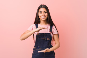 Wall Mural - Young pretty arab woman wearing a jeans dungaree holding something with both hands, product presentation.