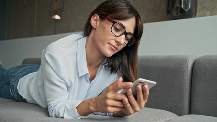 Wall Mural - Attractive calm young business woman resting and using smartphone while lying on sofa at home