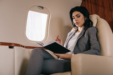 attractive businesswoman holding pen while looking at notebook in private jet