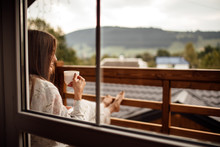 Young Woman On The Balcony Holding A Cup Of Coffee Ore Tea In The Morning. She In Hotel Room Looking At The Nature In Sumer. Girl Is Dressed In Stylish Nightwear. Relax Time.