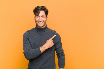 Wall Mural - young handsome man smiling cheerfully, feeling happy and pointing to the side and upwards, showing object in copy space against orange background