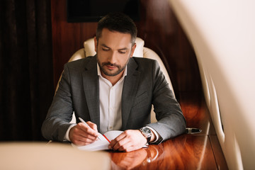 selective focus of bearded businessman holding pen while writing in notebook and sitting in plane